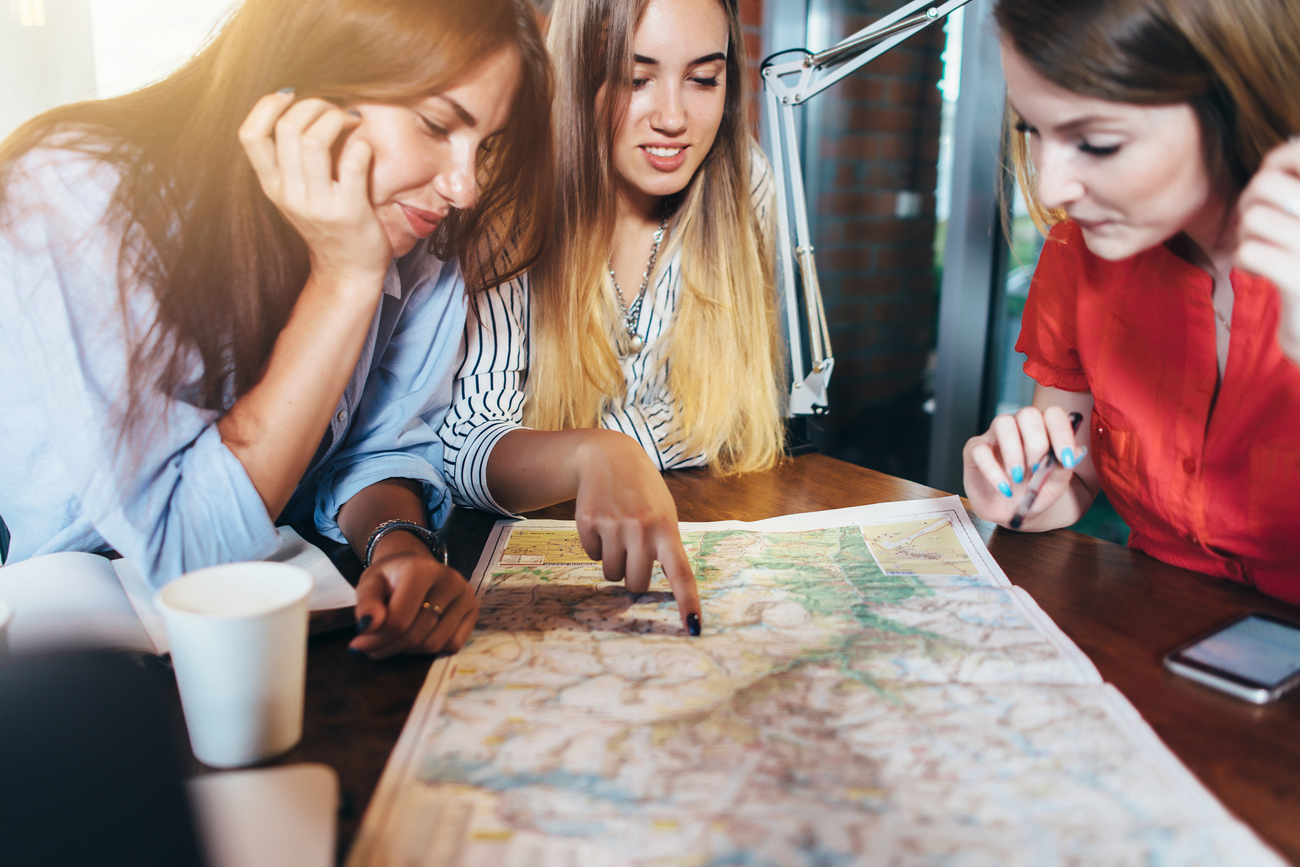 Three girlfriends planning their vacation sitting at table around map choosing the destination.
