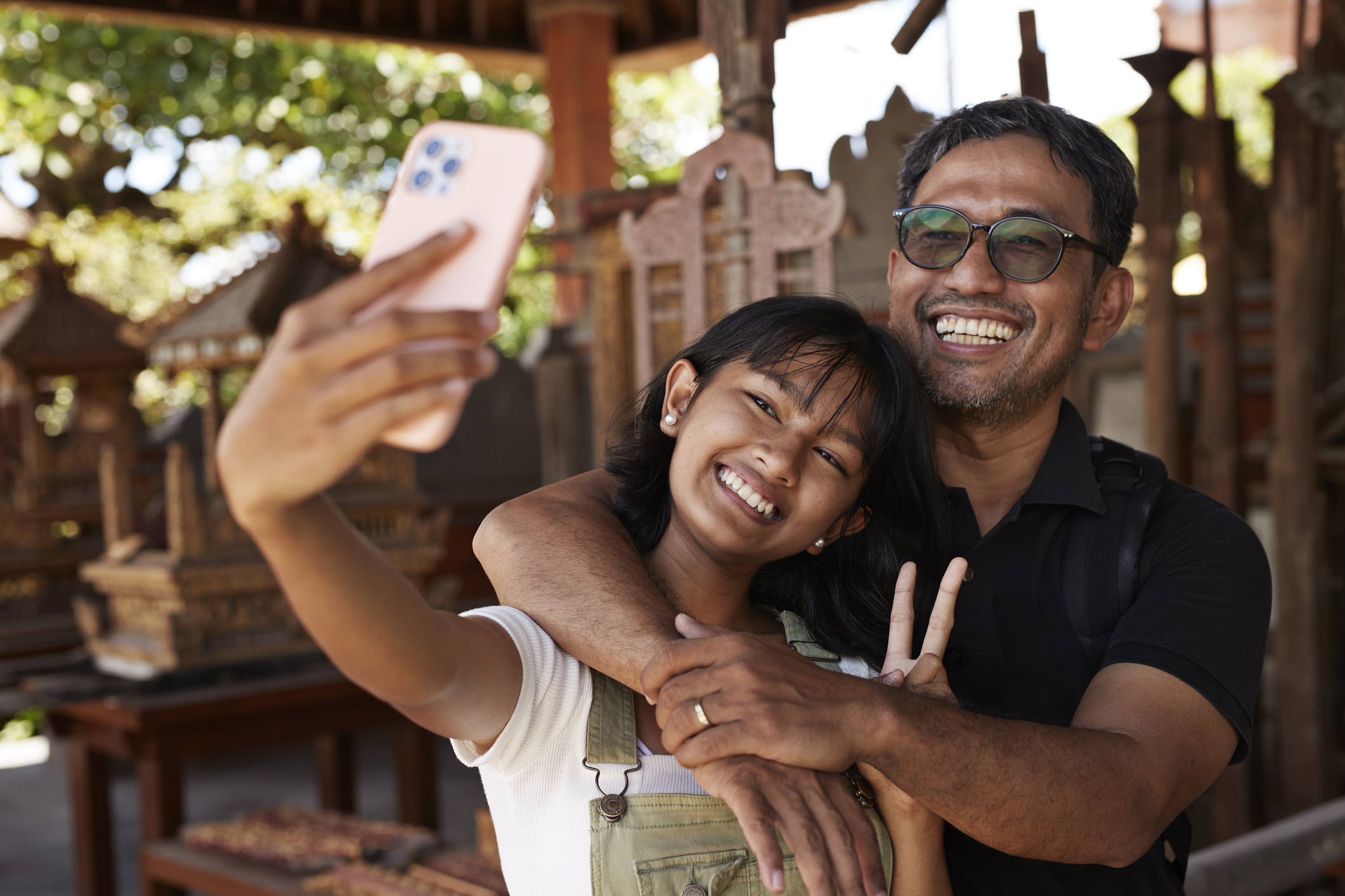 Happy girl showing peace sign while taking selfie with father through smart phone during vacation