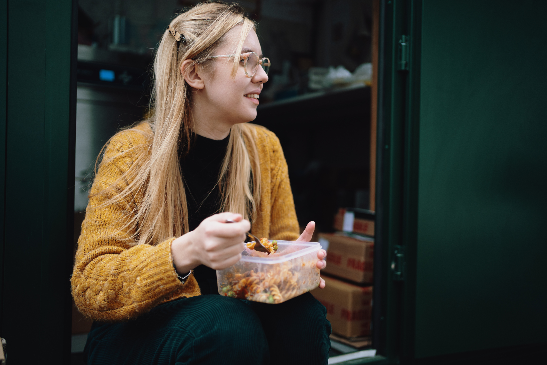 Woman with long blonde hair and glasses sat outside on step eating lunch from a lunchbox / Female Focus Collection