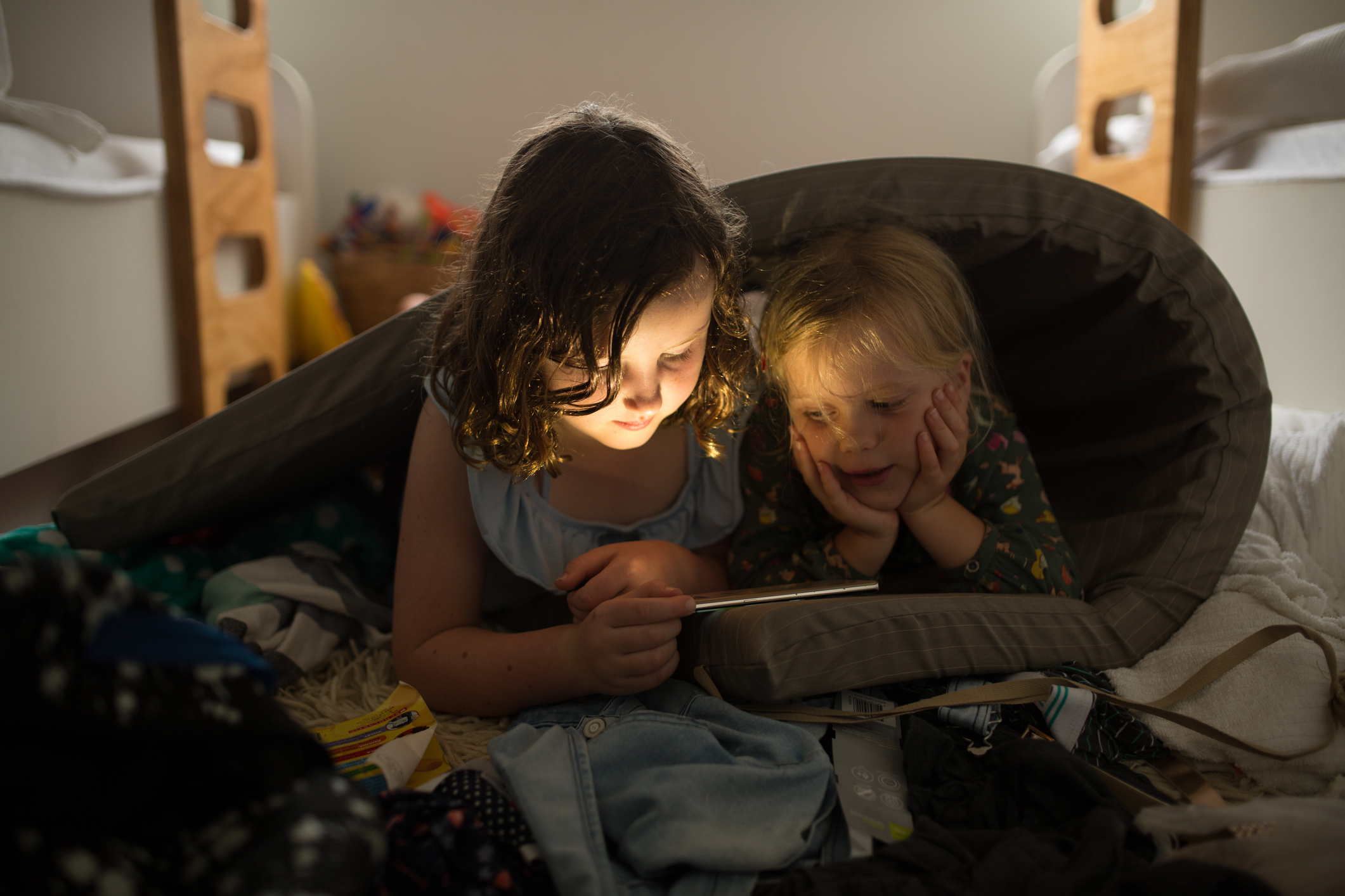 Two girls look at a digital tablet together wrapped in a mattress in a cluttered bedroom