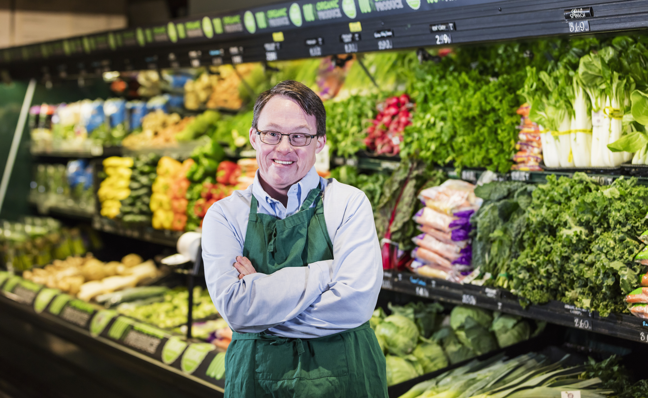 A mature man in his 40s with down syndrome working in a supermarket. He is wearing an apron, standing in the produce section of the store, looking toward the camera with his hands on his hips.
