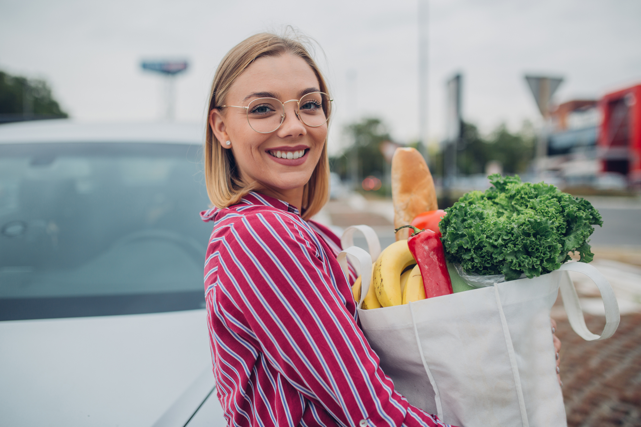 Young woman with eyeglasses standing with reusable bag on parking lot after groceries shopping