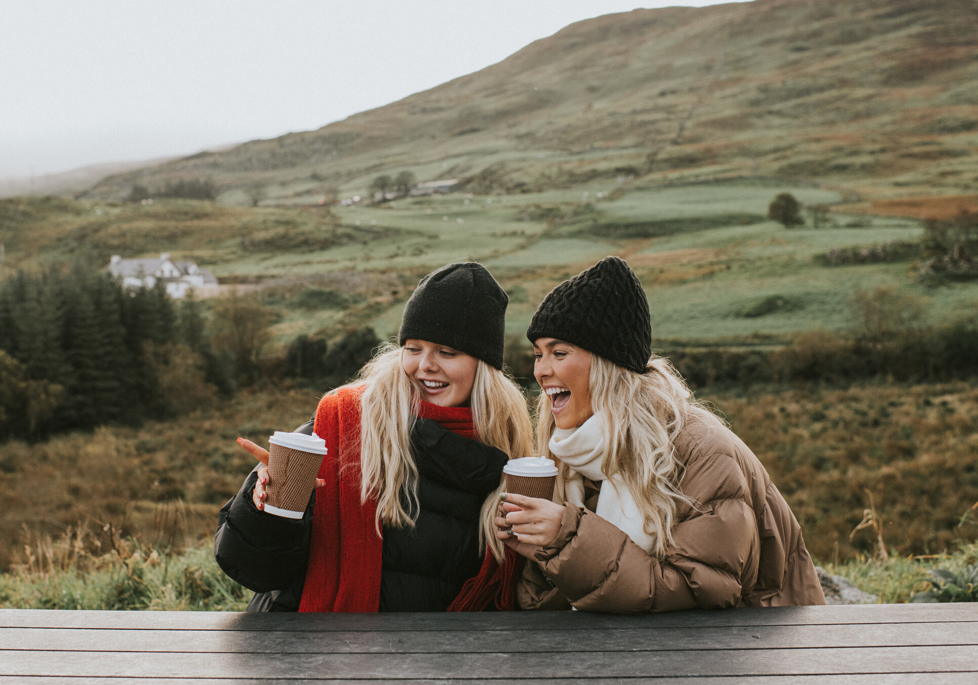 Two young woman holding takeaway coffee cups look cozy, wrapped up warmly in puffer jackets, hats and scarves, and snuggled up close to eachother. They smile and chat. One points at something.