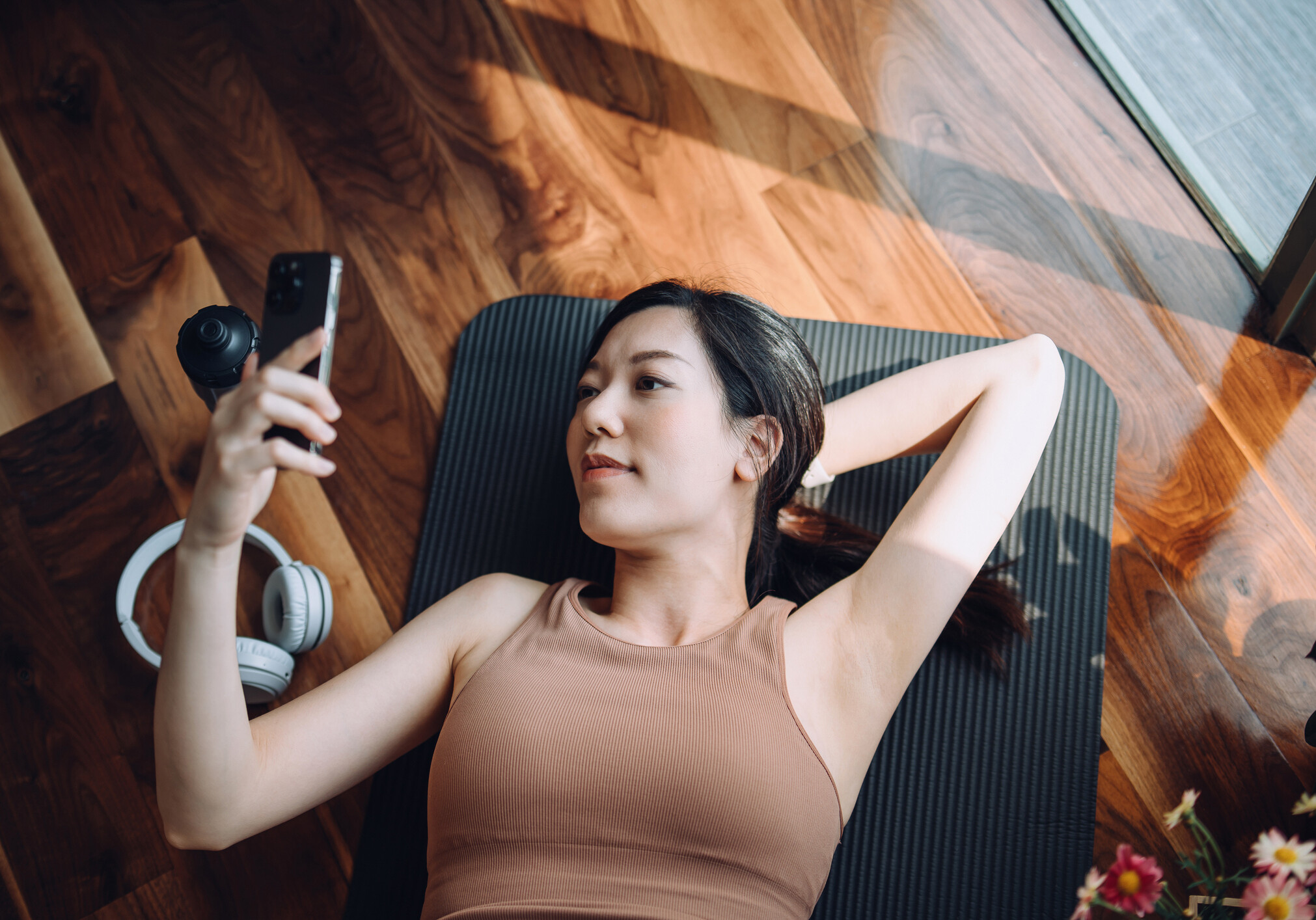High angle shot of fitness young Asian sports woman relaxing after working out, lying on yoga mat and using her smartphone against sunlight in the morning, reusable water bottle and headphones by her side. Health, fitness and wellness concept