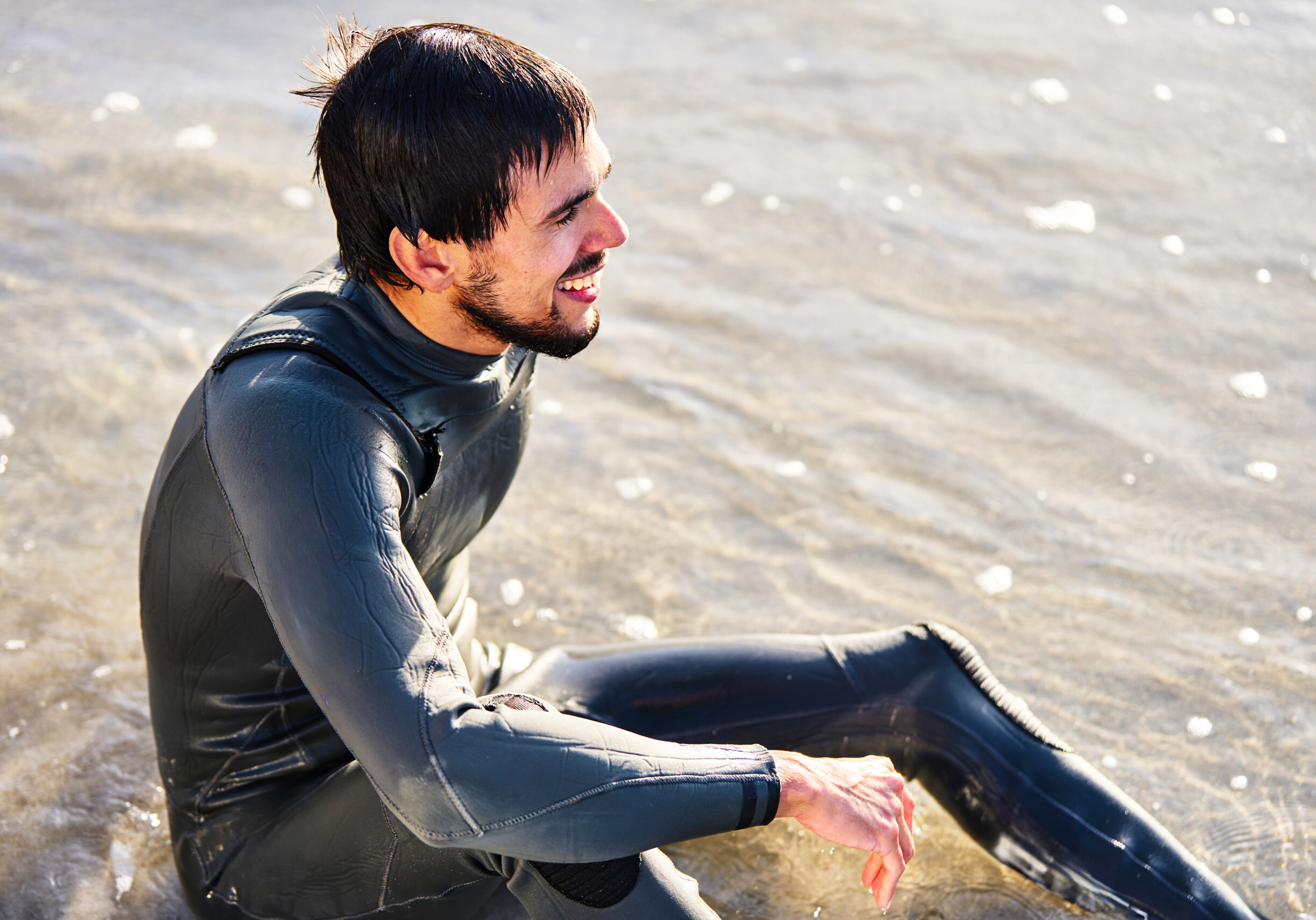A content male surfer in a wetsuit sits at the water's edge, basking in the sunlight with a pleased smile after an exhilarating surf session